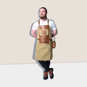 May include: A tan and brown apron with leather accents and pockets. The apron has a leather chest pocket and a leather holder for a bottle. The apron is worn by a person wearing a white t-shirt and black pants.