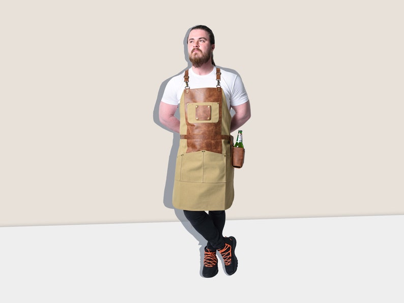 May include: A tan and brown apron with leather accents and pockets. The apron has a leather chest pocket and a leather holder for a bottle. The apron is worn by a person wearing a white t-shirt and black pants.