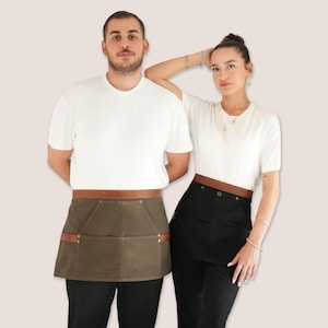 May include: Two people wearing white t-shirts and brown leather belts with brown and green canvas aprons. The person on the left is wearing a green apron with multiple pockets. The person on the right is wearing a black apron with two pockets.