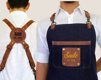 May include: A brown leather adjustable apron with a blue denim front panel. The apron has a large brown leather patch on the front with the text "Sade New York" in gold embroidery. The back of the apron has a brown leather patch with the text "Dad Est. 2022" in black.