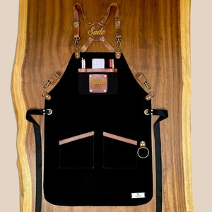 Black canvas apron with brown leather straps, custom embroidered name patch and front pockets