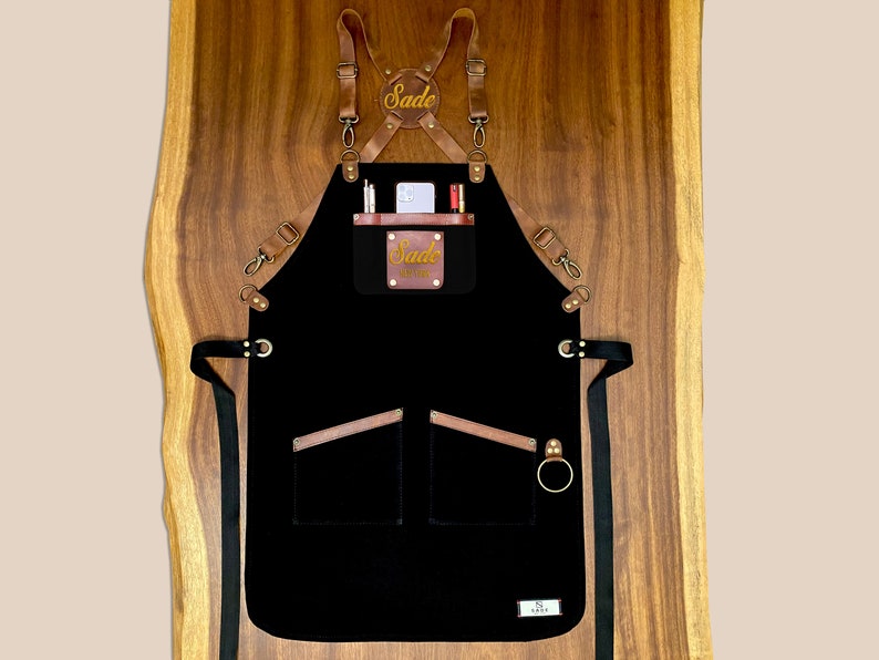 Black canvas apron with brown leather straps, custom embroidered name patch and front pockets