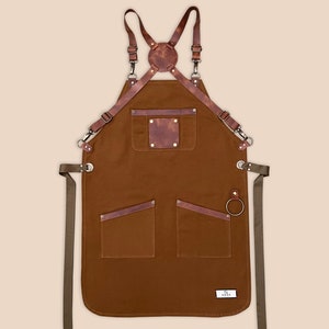 Brown canvas apron with adjustable leather cross-back straps, brass hardware and front pockets