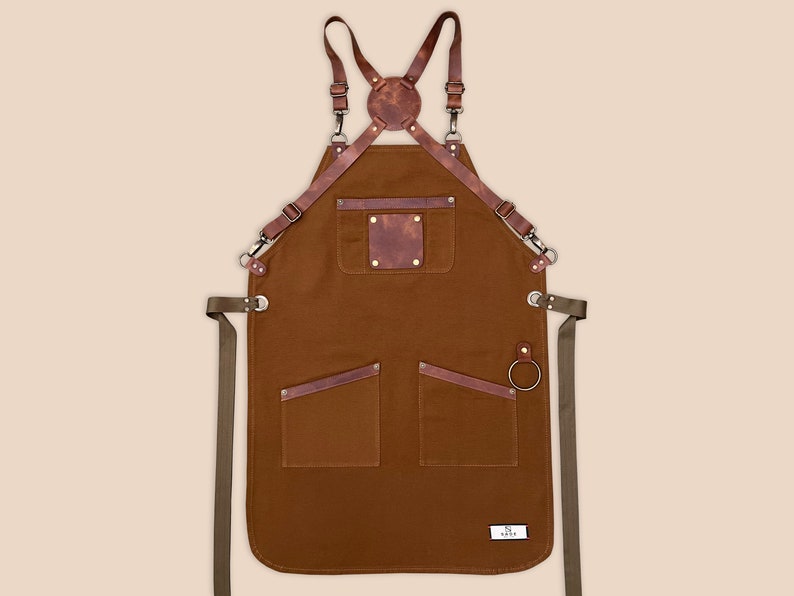 Brown canvas apron with adjustable leather cross-back straps, brass hardware and front pockets