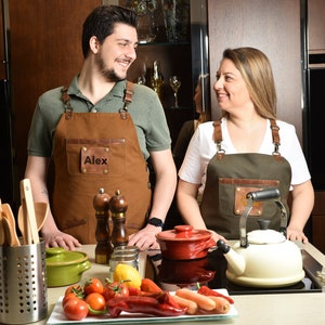 Two models wearing custom canvas aprons with leather accents in a modern kitchen setting
