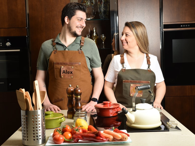 Two models wearing custom canvas aprons with leather accents in a modern kitchen setting