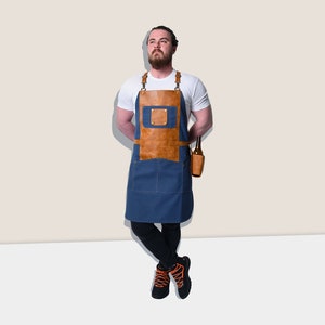May include: A blue denim apron with brown leather accents and straps. The apron has a front pocket and a leather beer bottle holder. The model is wearing a white t-shirt and black pants.