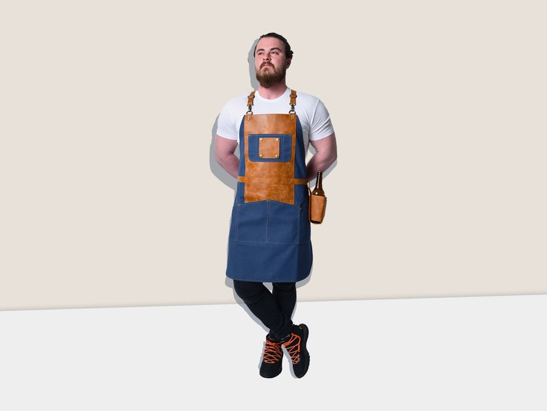 May include: A blue denim apron with brown leather accents and straps. The apron has a front pocket and a leather beer bottle holder. The model is wearing a white t-shirt and black pants.