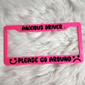 May include: Pink plastic license plate frame with black text that reads "Anxious Driver Please Go Around" with a smiley face and a sad face.