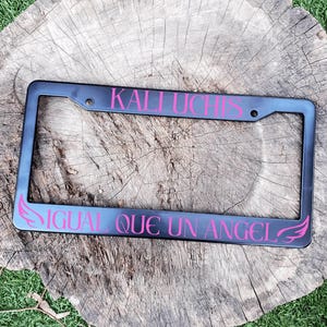 May include: Black metal license plate frame with pink text that reads "KALI UCHIS IGUAL QUE UN ANGEL" with angel wings on either side.
