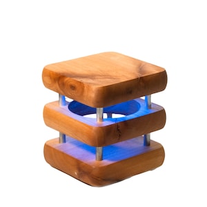May include: A wooden lamp with a blue light shining through the center. The lamp is made of three stacked square pieces of wood with a hole in the center.