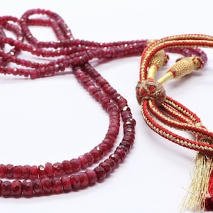 Puede incluir: Un collar de doble cadena hecho de cuentas de rubí rojo con un borla dorada y un cordón rojo y dorado.