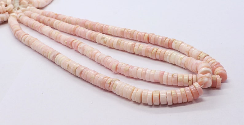Pode incluir: Close-up de v&aacute;rios fios de contas rosa e brancas em forma de disco. As contas s&atilde;o planas, circulares e t&ecirc;m uma superf&iacute;cie ligeiramente irregular. A cor &eacute; um rosa pastel suave com detalhes brancos.