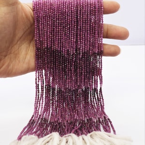 May include: A strand of pink ruby beads, approximately 2 mm in diameter, strung on a white thread. The beads are faceted and have a slight shimmer. The strand is about 18 inches long and is held in a hand.