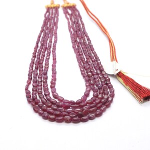Puede incluir: Un collar de varias hebras con cuentas ovaladas de color rojo intenso. Las cuentas están ensartadas, creando un efecto de capas. El collar tiene un cierre dorado y una borla roja y dorada.