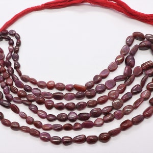 May include: A strand of deep red ruby gemstone beads, each bead is a smooth, oval shape. The beads are strung on a red cord with a tassel.