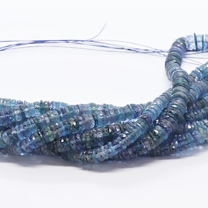 May include: A strand of blue gemstone beads, likely aquamarine, with a faceted, slightly irregular shape. The beads are strung on a thin wire and are arranged in a loose, flowing pile.
