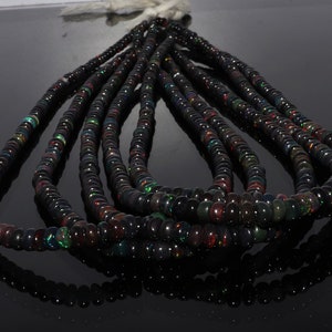 Puede incluir: Una hebra de cuentas de ópalo negro, con destellos iridiscentes de color, se muestra sobre una superficie negra. Las cuentas son redondas y lisas, y están dispuestas en un patrón suelto y fluido.