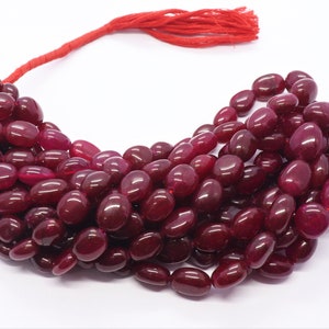 Può includere: Una collana di perle di pietra preziosa rosso scuro, ogni perla è di forma ovale e lucidata. Le perle sono infilate su un cordoncino rosso con un fiocco.