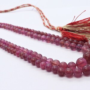 May include: A three-strand necklace made of pink round beads with a red and white tassel. The tag attached to the necklace reads "Gtu 296  Sz 28".