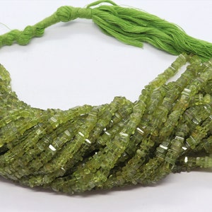 Könnte beinhalten: Eine Strähne grüner Peridot-Perlen, jede Perle ist quadratisch und facettiert. Die Perlen sind auf einer grünen Schnur mit einem grünen Quast aufgefädelt.