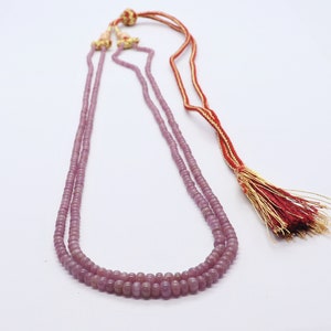 Puede incluir: Un collar de doble cadena hecho de cuentas de color púrpura claro con un borla roja y dorada.