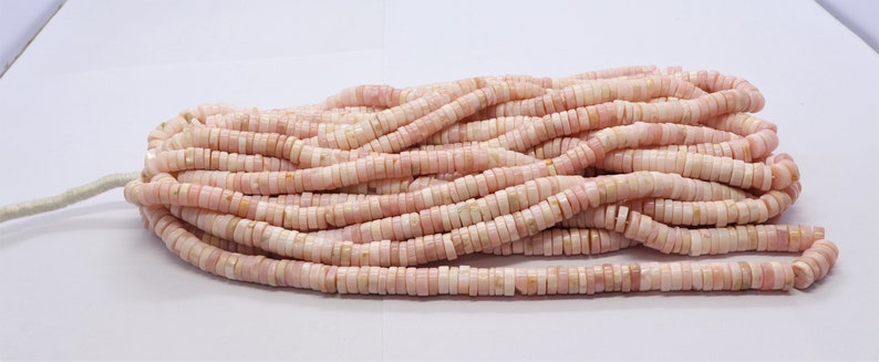 Pode incluir: Um fio de contas planas e redondas rosa e brancas. As contas s&atilde;o empilhadas, criando um conjunto espesso e texturizado. As contas s&atilde;o uma mistura de rosa p&aacute;lido e branco, com um leve brilho iridescente. As contas s&atilde;o enfiadas em um cord&atilde;o branco.