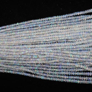 K&ouml;nnte beinhalten: Eine Str&auml;hne aus wei&szlig;en Opalperlen mit einem leichten irisierenden Schimmer. Die Perlen sind klein und rund und auf einem durchsichtigen Faden aufgef&auml;delt. Die Str&auml;hne liegt auf einer schwarzen Oberfl&auml;che.