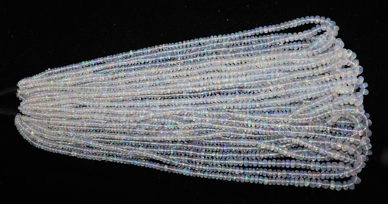 K&ouml;nnte beinhalten: Eine Str&auml;hne aus wei&szlig;en Opalperlen mit einem leichten irisierenden Schimmer. Die Perlen sind klein und rund und auf einem durchsichtigen Faden aufgef&auml;delt. Die Str&auml;hne liegt auf einer schwarzen Oberfl&auml;che.