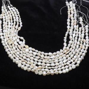 Puede incluir: Hebras de perlas blancas cremosas de forma irregular. Las perlas están ensartadas, creando múltiples hebras. El fondo es negro liso, lo que hace que las perlas destaquen. Estas perlas son adecuadas para la fabricación de joyas.