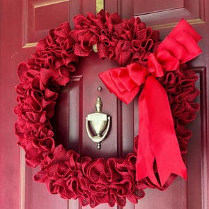 May include: A circular wreath constructed from ruffled red burlap fabric, featuring a large, smooth red satin ribbon bow attached to the right side. The wreath is mounted on a dark red door with a silver metal door knocker visible in the centre.