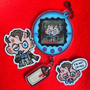 Puede incluir: Un conjunto de colgantes de acrílico y un llavero con un personaje de dibujos animados con un chupete. Los colgantes incluyen un biberón, un bocadillo de diálogo con el texto "I'M NOT CRYING!" y un dispositivo digital azul con un personaje.