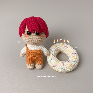 Puede incluir: Una muñeca de crochet con pelo rojo que lleva una camisa blanca y un mono naranja. La muñeca sostiene un donut de crochet blanco con chispas de colores.