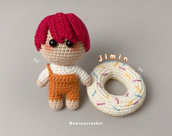 May include: A crocheted doll with red hair and a white shirt and orange overalls. The doll is holding a white crocheted donut with colorful sprinkles.
