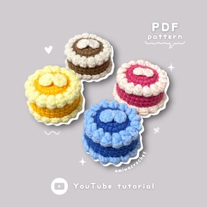 Puede incluir: Cuatro pasteles de crochet en diferentes colores: marrón, amarillo, rosa y azul. Cada pastel tiene un glaseado blanco y un diseño en forma de corazón en la parte superior. Los pasteles están dispuestos en círculo sobre un fondo gris. El texto "PDF pattern" y "YouTube tutorial" también es visible en la imagen.