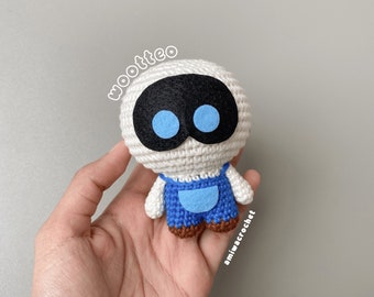 May include: White crocheted amigurumi toy with blue eyes and blue overalls. The toy is being held in a hand. The text "wootteo" and "amiwaccrochet" are visible in the image.