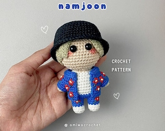 May include: Crocheted doll of a person wearing a black hat and a blue jacket with red flower details. The doll has a beige face and is labeled "namjoon" with "CROCHET PATTERN" text. The doll is held in a hand.