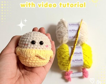 May include: A crochet kit featuring a small, round, cream and yellow banana-shaped plush toy with black eyes, alongside two skeins of yarn in cream and yellow, a gold crochet hook, and a small pink yarn tassel, with text reading CROCHET KIT.