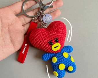 May include: Red heart-shaped crocheted keychain with a blue and yellow polka dot character attached. The keychain has a red velvet strap with the text "AMIWA.ID" and a silver charm with the text "AMIWA crochet".