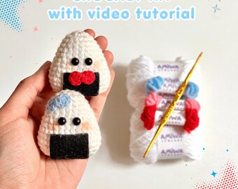 May include: A crochet kit featuring two white, triangular onigiri-shaped plushies with black felt bases, black bead eyes, and pink cheeks. One has a red bow, the other a blue bow. Includes white yarn skeins and a gold crochet hook. Text reads: CROCHET KIT with video tutorial.