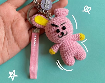 May include: Pink crocheted bunny keychain with yellow ears and black eyes. The keychain has a pink ribbon with the text "AMIWA.ID" and a small tag with the text "AMIWA CROCHET".