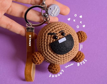 May include: Brown crocheted Shooky keychain with a black mouth and white teeth. The keychain has a brown velvet strap with the text "AMIWA ID" on it. The keychain is attached to a silver key ring with a purple charm that says "AMIWA crochet".