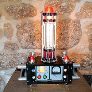 May include: An industrial-style lamp with a black base and a cylindrical, illuminated central column. The lamp has red accents and a red top, with the word "ATLAS" on the front. It sits on a wooden surface against a stone wall.