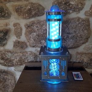 Pode incluir: Lâmpada futurista azul e prateada. Possui uma coluna central cilíndrica com luzes internas, uma base quadrada e um controle remoto. A lâmpada está sobre uma superfície de madeira contra uma parede de pedra. A lâmpada tem cerca de 30 cm de altura.