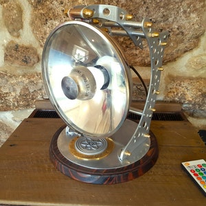 May include: A steampunk-style desk lamp with a large, reflective, circular light. The lamp features a silver metal frame, a wooden base, and a remote control with color options. The light source is labeled "URANA OHIO ST-124".