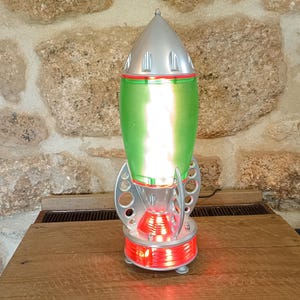 May include: A retro rocket lamp with a silver top, green body, and red accents. The lamp has a circular base and fins, and it is illuminated with red and white lights. The lamp is approximately 30 cm tall.