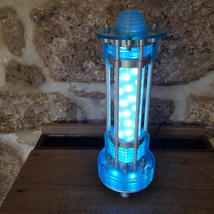 May include: A futuristic-looking lamp with a cylindrical design. The lamp features a blue, transparent body with internal LED lights, and silver metal accents. The top has a dome-shaped cover. The lamp is on a wooden surface.