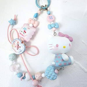 May include: A pastel-colored keychain featuring a Hello Kitty figure, bows, stars, and beads. The keychain includes a blue clasp and a variety of decorative elements in pink, blue, and white. The Hello Kitty figure is wearing a blue dress with pink hearts.