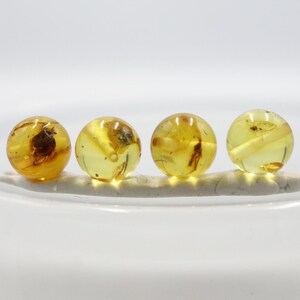 May include: Four spherical amber beads with inclusions, displayed on a light-coloured, oblong base. The beads are a translucent yellow-orange colour, set against a white background.