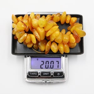 May include: A necklace of polished amber beads, in shades of yellow and orange, sits on a black tray on a digital scale. The scale shows a weight of 20.07 grams. The beads are irregularly shaped and strung together.